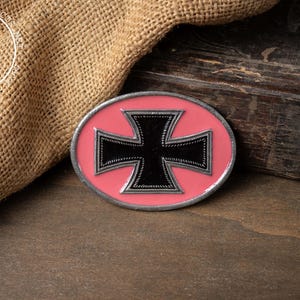 May include: An oval-shaped metal belt buckle featuring a black cross emblem centered on a solid pink background. The buckle has a silver-toned metallic border and a raised, textured edge surrounding the black cross design, with a circular logo in the corner.