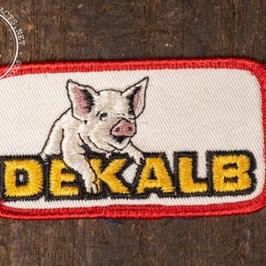 May include: Embroidered patch featuring a pig illustration above the word "DEKALB" in yellow and black. The patch has a white background with a red border, and is likely a vintage collectible or souvenir.