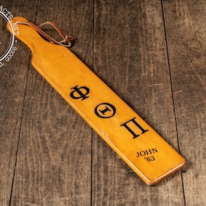 May include: A wooden paddle with a light brown finish, featuring the Greek letters Phi, Theta, and Pi in black. The name "John '63" is also inscribed. A leather loop is attached to the handle.