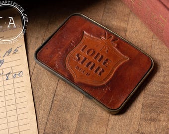 May include: A brown leather belt buckle with a brass frame. The buckle features a raised, embossed logo of a shield with a star and the words "Lone Star Beer".