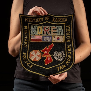 May include: A black felt patch with gold embroidery. The patch is shaped like a shield and features the words "Memory of Korea" and "Korea" in large letters. The patch also includes several smaller embroidered patches, including a US flag, a Korean flag, and a UN flag.