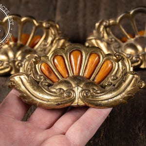 May include: Ornate, antique brass drawer pulls with a fan-shaped design and amber-colored accents. The vintage hardware features detailed scrollwork and a warm, aged patina, perfect for adding a touch of elegance to furniture.