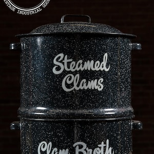 May include: Black speckled enamel clam steamer with the words "Steamed Clams" and "Clam Broth" in white script. The steamer has a lid, handles, and a spigot. The background is a dark wood.