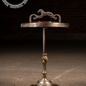 May include: Vintage metal ashtray stand with a circular tray and a decorative horse silhouette. The stand features a detailed base and a central pole, showcasing an antique design. The ashtray is made of metal and stands approximately 30 inches tall.