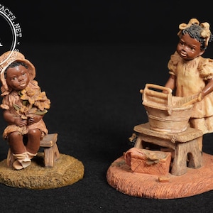 May include: Two vintage figurines depicting African American girls. One girl sits holding flowers, the other stands by a wash basin. Both are dressed in light-colored dresses and are on small stools. The figurines are on a brown and tan base.