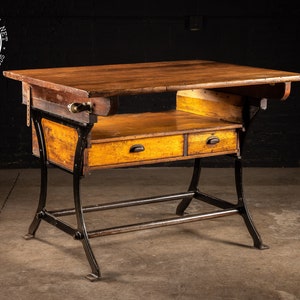 May include: A vintage industrial work table with a wooden top and two drawers. The table has a black metal frame with a lower shelf. The table is in good condition and has a rustic look.