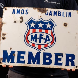 May include: White metal sign with blue and red text and a red, white, and blue shield with three stars and the letters "M-F-A". The sign reads "Amos Gamblin Member".
