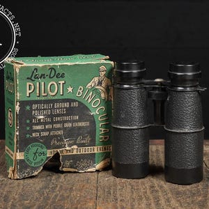 May include: Vintage Lan-Dee Pilot binoculars with a green and black box. The binoculars are black with a textured grip. The box features text that reads "Pilot Binocular" and "Optically Ground and Polished Lenses."