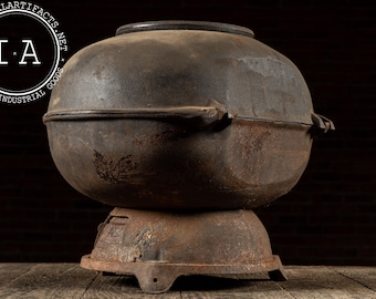 May include: An antique, cast iron cooking pot with a rounded body and a separate base. The pot is dark brown with a rusted appearance, suggesting age and use. The pot has a lid and handles. The base has a decorative design.
