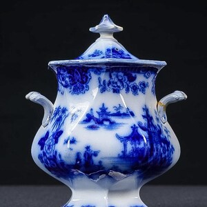 May include: A blue and white porcelain sugar bowl with a lid. The bowl features a detailed landscape scene with figures and buildings. The handles are curved and the lid has a finial. The design is a classic blue and white pattern.