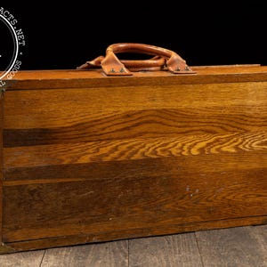 May include: A vintage wooden box with a leather handle and brass corner protectors. The box has a rich, warm wood grain pattern and is likely used for storage or display. The box is rectangular and sits on a wooden surface.
