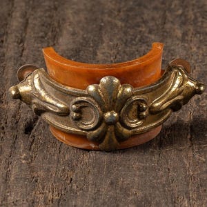 May include: Three antique brass and leather drawer pulls. Each pull features a curved, orange-toned leather base with ornate brass detailing. The pulls are arranged on a weathered wooden surface, showcasing their vintage aesthetic.