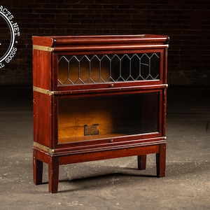 May include: A vintage wooden bookcase with two sections, featuring glass doors and brass accents. The top section has a decorative leaded glass design. The bookcase is a rich, dark brown color and stands on four short legs. The logo "Industrial Artifacts" is in the top left corner.