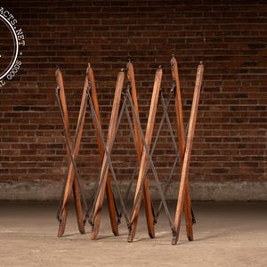 May include: A folding wooden luggage rack featuring a brown finish and a crisscross accordion-style frame. The structure is composed of vertical wooden slats connected by dark metal hardware, allowing the unit to expand and contract.