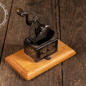 May include: An antique pencil sharpener with a wooden handle and a metal body, mounted on a wooden base. The sharpener is black with some visible text and is set against a rustic wooden background.