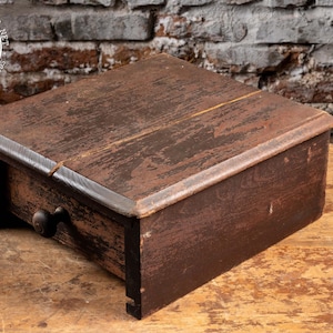 May include: A small, antique wooden box with a drawer. The box is stained a dark brown with visible wood grain and wear. It has a simple, rectangular design with a small, round drawer pull. The box sits on a wooden surface.