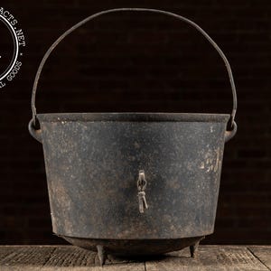 May include: A weathered, black cast iron cauldron with a curved metal handle. The cauldron has three small legs and a spigot. The image includes the text "INDUSTRIALARTIFACTS.NET" and "VINTAGE INDUSTRIAL GOODS".