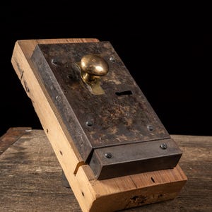 May include: Antique metal door lock with a brass knob and keyhole, mounted on a wooden base. The lock has a weathered, aged appearance, with visible screws and a dark, textured metal surface. The wooden base is made of light-colored wood.
