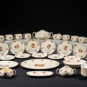 May include: A large cream-colored ceramic dinnerware set featuring a central floral motif with blue, yellow, and pink petals. The collection includes numerous plates, bowls, cups, a lidded tureen, and a gravy boat, all arranged on a dark surface.