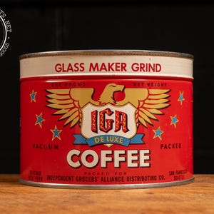 May include: Vintage IGA De Luxe Coffee tin. The red tin features a gold eagle emblem, blue stars, and the words "Glass Maker Grind" and "Coffee." The tin is marked "One Pound" and "Packed for Independent Grocers' Alliance Distributing Co."