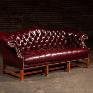 May include: A burgundy leather Chesterfield sofa with button tufting and wooden legs. The sofa has rolled arms and a low back. The frame is made of wood and the cushions are leather. The sofa is a classic piece of furniture.