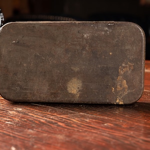 May include: A vintage metal tin box with a worn, brown surface. The box is rectangular and has a slightly rounded edge. The surface of the box is covered in scratches and scuffs, giving it a rustic look.