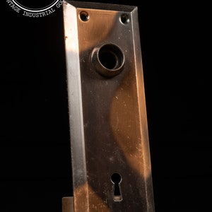 May include: A vintage brass door plate with a keyhole and a round knob. The plate has a worn, aged look with a dark brown patina.