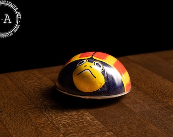 May include: A vintage tin toy ladybug with a yellow face and black eyes. The ladybug is red, yellow, and blue.