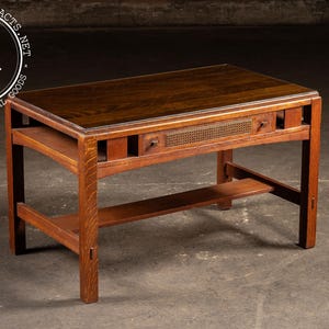 May include: A rectangular wooden desk with a dark brown top and a lighter brown frame. The desk features a drawer with a woven panel and small pulls. A lower shelf and sturdy legs complete the design. The logo "I.A" is in a circle in the upper left corner.