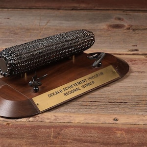 May include: A bronze-colored corn sculpture mounted on a wooden base. The base has a brass plaque that reads "DEKALB ACHIEVEMENT PROGRAM REGIONAL WINNER". The sculpture is a unique award or decorative piece.