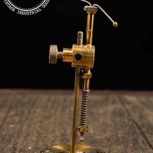 May include: Vintage brass Keleket tool with a spring and adjustable arm. The tool has a rectangular base and a cylindrical body with a screw for adjustments. The image is set against a dark background, highlighting the tool's details.
