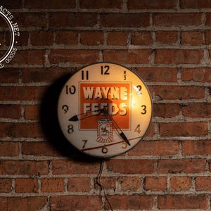 May include: Vintage Wayne Feeds clock with a white face, black hands, and orange lettering against a brick wall. The clock features the text "WAYNE FEEDS" and a logo. The clock is illuminated and has a power cord.