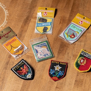 May include: A collection of vintage embroidered patches featuring various states and locations, including Nebraska, Illinois, Iowa, Värmland, Estes Park, Colorado, and Portugal. The patches are yellow, blue, green, red, and white with embroidered details.