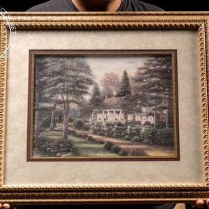 May include: A framed print of a painting depicting a white cottage with a green roof surrounded by trees and a path leading to the front door. The painting is set against a backdrop of a blue sky with white clouds. The frame is gold with a decorative border.