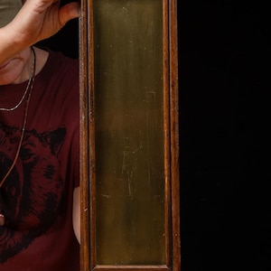 May include: A tall, rectangular wall-mounted object featuring a dark brown wooden frame surrounding a smooth, vertical brass panel. The top of the frame has a rounded arch with a circular hole, and a small brass plumb bob hangs from the bottom.
