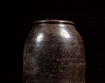 May include: A dark brown, antique stoneware crock with a rounded body and a slightly flared rim. The surface has a mottled appearance, and the piece sits on a wooden surface. The image includes the text "Vintage Industrial Goods".