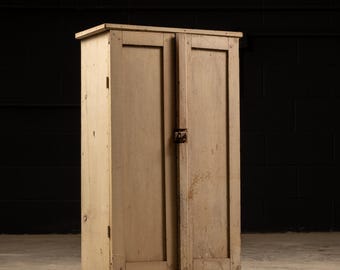 May include: A vintage wooden cabinet with two doors and a simple design. The cabinet is painted a light beige color and has a slightly distressed look. The cabinet is standing on a concrete floor.