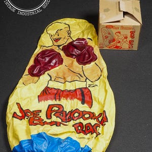 May include: Vintage inflatable "Joe Palooka" bop bag. The yellow bag features a cartoon boxer with red gloves and shorts. The text "Joe Palooka Bop Bag" is printed in red. A cardboard box with the same design is also present.