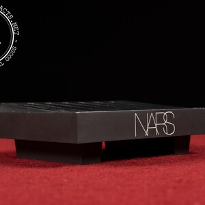 May include: A black rectangular display stand with the word "NARS" in silver lettering. The stand is on a red surface with a black background. The stand has a layered design.