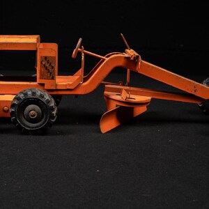 May include: A vintage orange metal toy road grader featuring a central blade, a steering wheel, and six black rubber tires with deep treads. The side of the frame displays a small, rectangular black and orange logo with the text Tonka Toys.