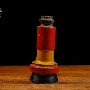 May include: A vintage, hand-painted wooden bottle stopper. The stopper features a cylindrical design with sections of red, yellow, and black paint. The top is a dark, weathered color. The item is displayed on a wooden surface against a black background.