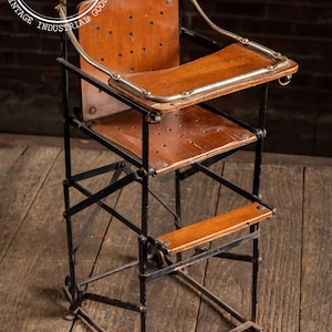Puede incluir: Silla alta vintage con asiento y bandeja de madera, y estructura de metal negro. La silla tiene reposapiés y respaldo. El asiento y la bandeja son de madera. La silla es plegable.