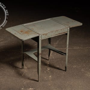 May include: Vintage industrial metal folding table in a muted green color. The table has a rectangular top with hinged leaves and a lower shelf. The metal legs are slender and angled, with visible rust spots adding character. The table is designed to fold for storage.