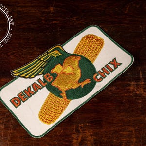 May include: A rectangular embroidered patch with a white background and a green border. The design features a yellow ear of corn, a green circle containing a yellow chick, a yellow wing, and the orange text DEKALB CHIX printed on the white fabric.