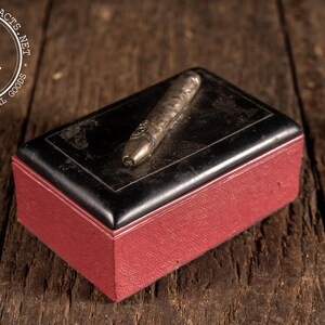 May include: A rectangular box with a deep red textured base and a smooth black lid featuring a thin white border. Resting on top of the lid is a metallic, cylindrical object with a tapered end and a small circular emblem on its surface.