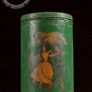 May include: Vintage green metal tin with a cylindrical shape. The tin features a colorful illustration of people dancing, with a woman in a yellow dress. The tin has a distressed appearance, with the text "INDUSTRIAL ARTIFACTS" and "I A" on the top.