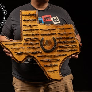 May include: A wooden Texas-shaped plaque with a horseshoe and various barbed wire samples mounted on it. The plaque is held by a person wearing a dark grey t-shirt and khaki pants.