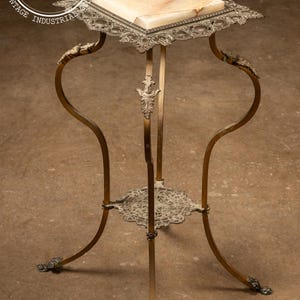 May include: A gold metal stand with a white marble top and ornate details. The stand has three legs with claw feet and a lower shelf.