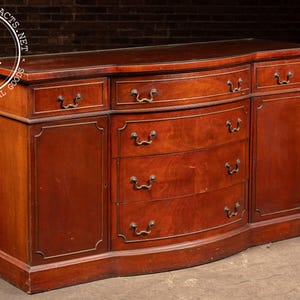 May include: A mahogany sideboard featuring a curved front, three top drawers, three central drawers, and two side cabinets. The piece includes dark metal hardware pulls and a watermark in the top left corner reading INDUSTRIALARTIFACTS.NET I A.