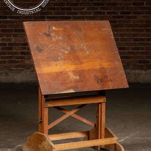 May include: A vintage wooden drafting table with a slanted top and adjustable height. The table is made of light brown wood and has a sturdy base. The image includes the text "Vintage Industrial Goods".